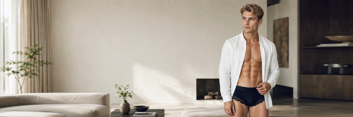 Man wearing dark blue mid-rise briefs and an unbuttoned white shirt standing near a window.