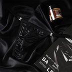 A lifestyle flat lay photograph featuring the MemoSkin Black Satin Low-Rise Men's Thong with adjustable metal buckle side straps, set on luxurious dark silk fabric. Crafted with smooth fabric and flexible side adjustments for personalized comfort, this minimalist men's thong underwear combines sleek aesthetics with everyday breathable comfort for modern men's wear.