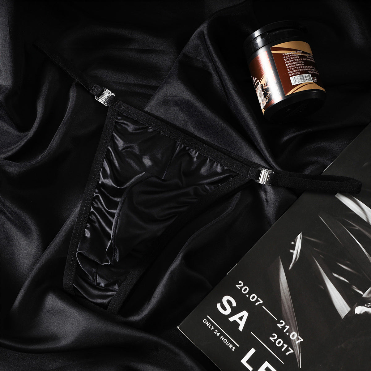 A lifestyle flat lay photograph featuring the MemoSkin Black Satin Low-Rise Men's Thong with adjustable metal buckle side straps, set on luxurious dark silk fabric. Crafted with smooth fabric and flexible side adjustments for personalized comfort, this minimalist men's thong underwear combines sleek aesthetics with everyday breathable comfort for modern men's wear.